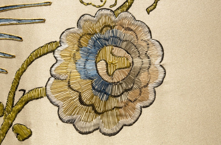 Fragment haftu na tkaninie. Jest to kwiat wyszywany złotymi, srebrnymi i błękitnymi nićmi. 