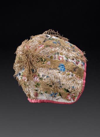 Zdjęcie haftowanego czepca. Na jasnym jedwabiu kolorowe hafty w kształcie kwiatów. Całość pokryta warstwą siatki złotych haftów.  